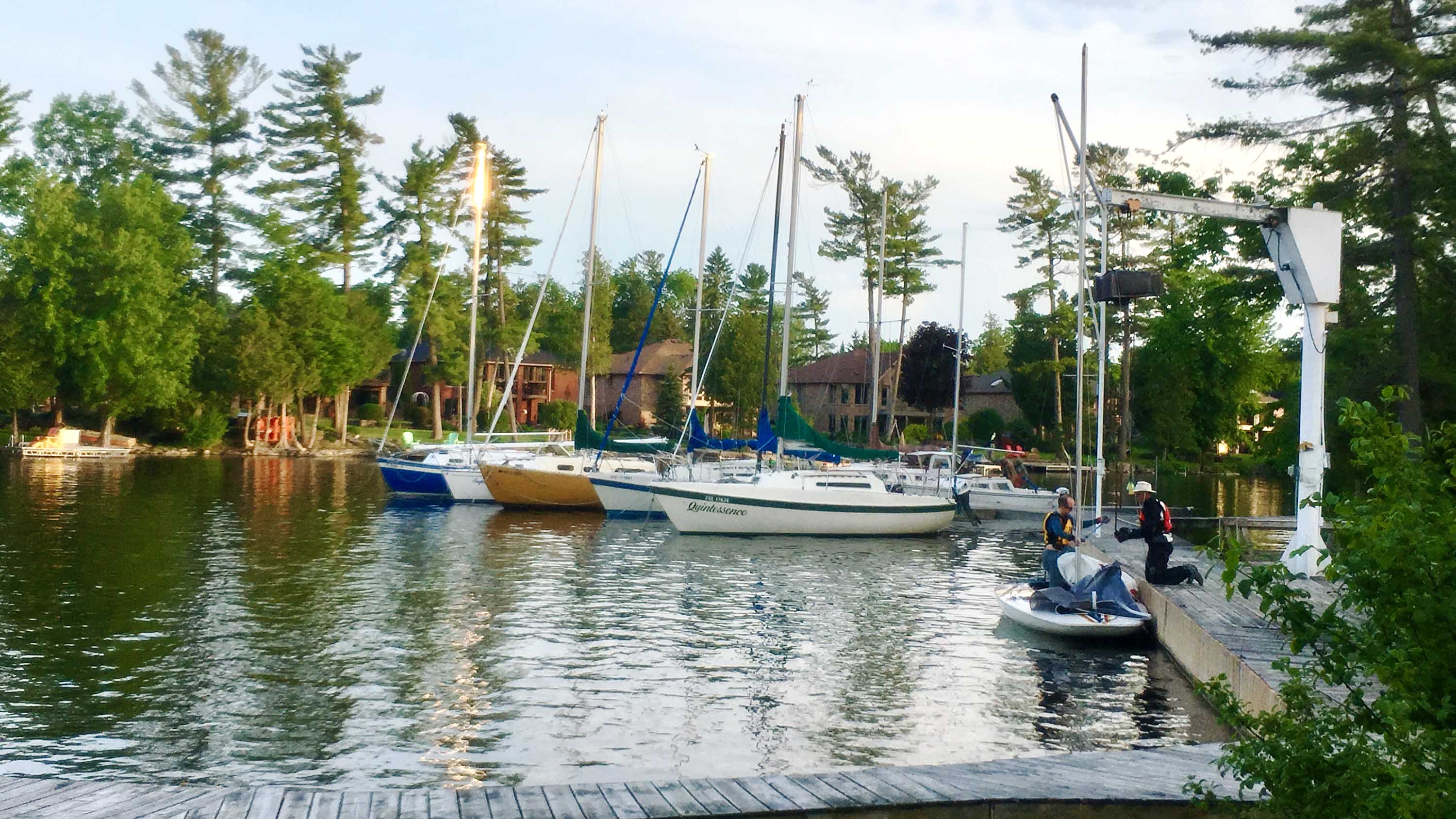 Photo of docks and several sailboats. There are two men preparing to lift a small sailboat out of the water using a hoist.